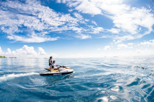 A person rides a jet ski on a vast ocean under a vibrant blue sky dotted with clouds. The deep blue water glistens as they speed along, while the distant shoreline with trees is visible on the horizon, painting a serene scene far from any need for a Tampa Boat Accident Lawyer.