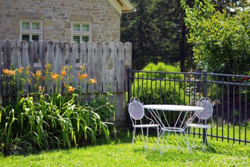 A quaint garden scene with a white metal table and two chairs on a grassy lawn, where bright orange daylilies bloom beside a rustic wooden fence. A stone house stands in the background, bordered by lush greenery and a metal fence that encloses this serene escape shared with my friend.