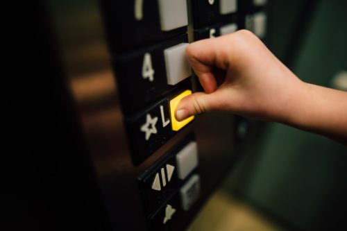 A close-up of a child's hand cautiously pressing the "L" button in an elevator. The illuminated button is surrounded by darkened ones, each bearing witness to countless journeys without incident. The metallic control panel with raised numbers stands resilient against any chance of an elevator accident.